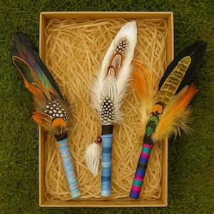 May include: Three decorative feather wands arranged in a rectangular box filled with shredded paper. Each wand features a unique combination of natural bird feathers in shades of white, black, orange, and green, attached to handles wrapped in colourful thread.