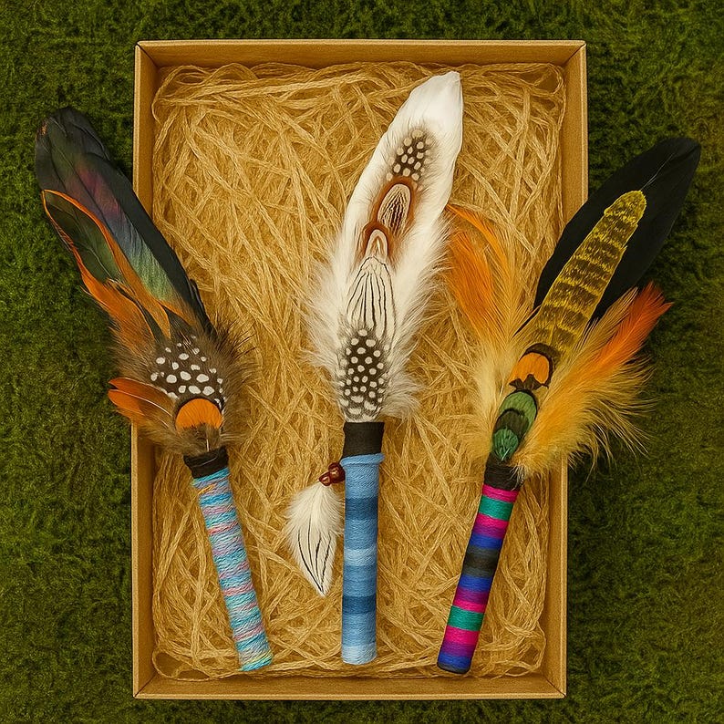 May include: Three decorative feather wands arranged in a rectangular box filled with shredded paper. Each wand features a unique combination of natural bird feathers in shades of white, black, orange, and green, attached to handles wrapped in colourful thread.