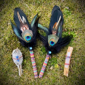 May include: Two ceremonial fans crafted with black feathers and iridescent peacock eye feathers, featuring handles wrapped in multicoloured thread. Beside them are a bundle of dried white sage and a rectangular piece of palo santo wood.