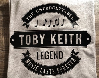 May include: Gray t-shirt with a black graphic design featuring the text "The Unforgettable Toby Keith Legend Music Lasts Forever" with musical notes and a banner.
