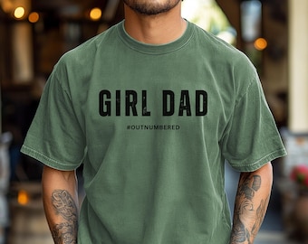 May include: A moss green t-shirt with the text "GIRL DAD" in bold black letters, with "#OUTNUMBERED" below. The shirt is a short-sleeved, crew neck style.