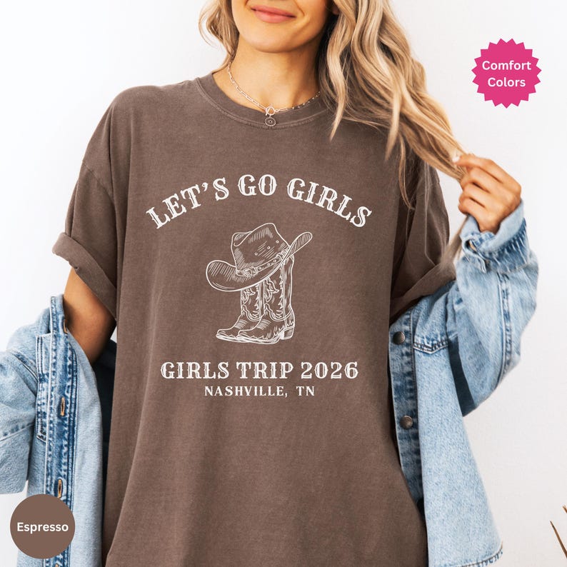 May include: Espresso-colored t-shirt with the text "LET'S GO GIRLS" above a graphic of a cowboy hat and boots. Below the graphic, it reads "GIRLS TRIP 2026 NASHVILLE, TN". The shirt has rolled-up sleeves and a relaxed fit.