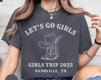 May include: Dark grey t-shirt featuring the text "LET'S GO GIRLS" above a cowboy hat and boots graphic. Below the graphic, the text reads "GIRLS TRIP 2025 NASHVILLE, TN". The shirt is a casual style.