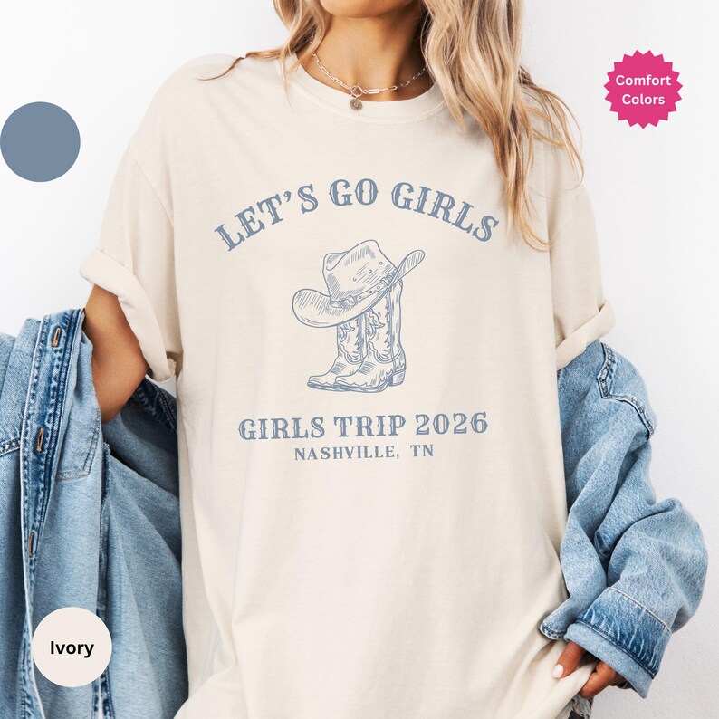 May include: Ivory-colored t-shirt with the text "LET'S GO GIRLS" above a graphic of a cowboy hat and boots. Below the graphic is "GIRLS TRIP 2026 NASHVILLE, TN". The shirt has a relaxed fit and rolled sleeves.