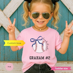 May include: Pink toddler-sized t-shirt with a baseball graphic and the text "baseball sister" and "GRAHAM #2". The baseball graphic includes a blue bow. The girl is wearing sunglasses and making a peace sign.