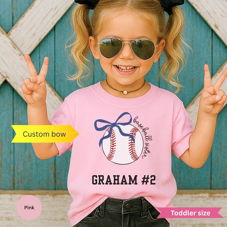 May include: Pink toddler-sized t-shirt with a baseball graphic and the text "baseball sister" and "GRAHAM #2". The baseball graphic includes a blue bow. The girl is wearing sunglasses and making a peace sign.