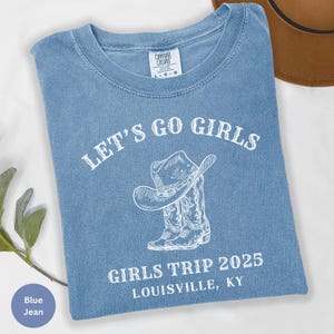 May include: A blue jean t-shirt with the text "LET'S GO GIRLS" above a graphic of a cowboy hat and boots. Below the graphic, the text reads "GIRLS TRIP 2025 LOUISVILLE, KY". The shirt has a crew neck and short sleeves.