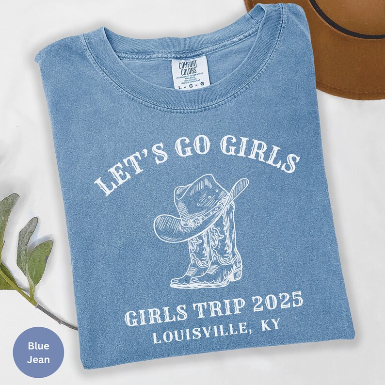 May include: A blue jean t-shirt with the text "LET'S GO GIRLS" above a graphic of a cowboy hat and boots. Below the graphic, the text reads "GIRLS TRIP 2025 LOUISVILLE, KY". The shirt has a crew neck and short sleeves.