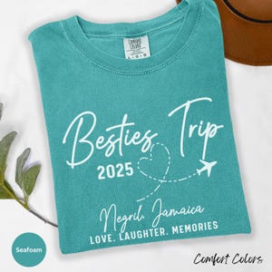 May include: A seafoam blue t-shirt with white text that reads "Besties Trip 2025" with a heart and airplane graphic. The text "Negril Jamaica" and "LOVE. LAUGHTER. MEMORIES" are also printed on the shirt.