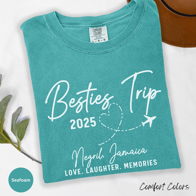 May include: A seafoam blue t-shirt with white text that reads "Besties Trip 2025" with a heart and airplane graphic. The text "Negril Jamaica" and "LOVE. LAUGHTER. MEMORIES" are also printed on the shirt.