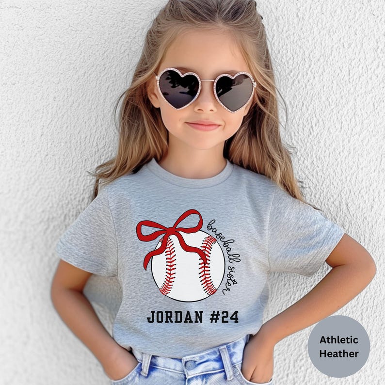 May include: A light gray athletic heather t-shirt featuring a baseball design with a red bow and the words "baseball sister." The shirt also includes the name "JORDAN" and the number "24" in black text.