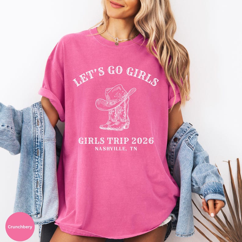 May include: A pink t-shirt with the text "LET'S GO GIRLS" above a graphic of a cowboy hat and boots. Below the graphic is "GIRLS TRIP 2026 NASHVILLE, TN". The shirt is worn with a denim jacket.