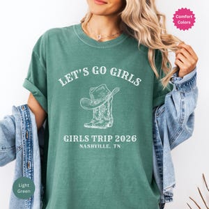 May include: Light green t-shirt with the text "LET'S GO GIRLS" above a graphic of a cowboy hat and boots. Below the graphic is "GIRLS TRIP 2026 NASHVILLE, TN". The shirt has rolled-up sleeves.