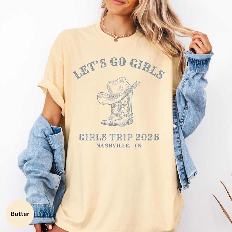 May include: Butter-colored t-shirt with the text "LET'S GO GIRLS" above a graphic of a cowboy hat and boots. Below the graphic, the text reads "GIRLS TRIP 2026 NASHVILLE, TN". The shirt has short sleeves and a relaxed fit.