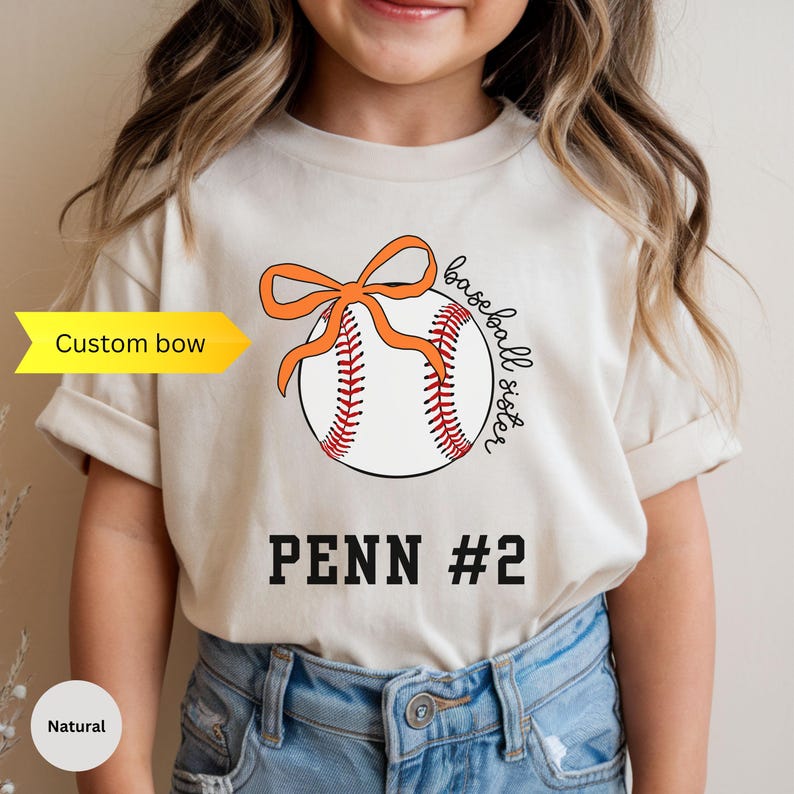May include: A natural-colored t-shirt featuring a baseball design with an orange bow and the words "baseball sister." Below the baseball is the text "PENN #2." The shirt has short sleeves and is worn by a child.