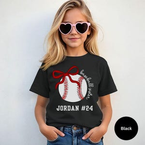 May include: Black t-shirt featuring a baseball design with a red bow and stitching. The text "baseball sister" is written in cursive, with the name "JORDAN #24" below the baseball. The shirt is worn by a person wearing pink heart-shaped sunglasses.