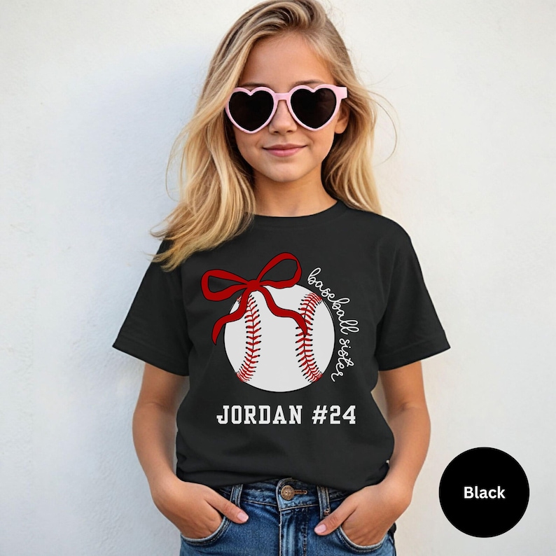 May include: Black t-shirt featuring a baseball design with a red bow and stitching. The text "baseball sister" is written in cursive, with the name "JORDAN #24" below the baseball. The shirt is worn by a person wearing pink heart-shaped sunglasses.