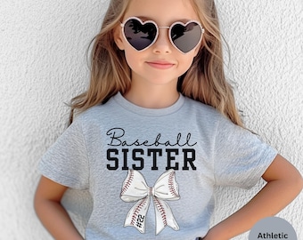 May include: A gray t-shirt with the text "Baseball Sister" and a white bow with red stitching and a baseball design. The bow has the number "#22" on it. The shirt is on a young girl wearing heart-shaped sunglasses.
