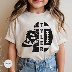 May include: A cream-colored short-sleeve t-shirt featuring a black graphic of a football player in a helmet, the name TYLER written vertically, and a football. The player's jersey displays the number 87. The shirt is paired with blue denim jeans.