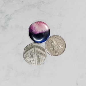 May include: A round button with a pink and purple gradient design featuring a white line and a dark forest silhouette. The button is next to two British coins.
