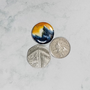 May include: A small, round button featuring a painted image of a mountain scene. The scene depicts a yellow sunset, a mountain peak, and a dark blue sky with a flock of birds flying overhead.
