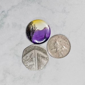 May include: A purple, yellow and black button with a silhouette of a tree and mountains. The button is next to two British coins.