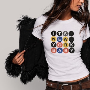 Puede incluir: Una camiseta blanca de manga corta con un estampado gr&aacute;fico de cuatro filas de c&iacute;rculos. Los c&iacute;rculos contienen letras que forman la frase ITS NEW YORK BABY en colores negro, amarillo, rojo y azul, imitando la est&eacute;tica de un letrero de metro.