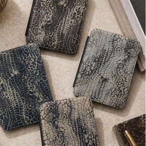 May include: Four rectangular, leather-like wallets with a reptile skin pattern in shades of gray, blue, and beige. Each wallet has a black spine. The wallets are arranged on a neutral surface, suggesting a collection or display of accessories.