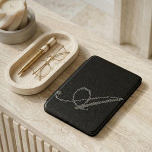 May include: A black tablet case with a stitched design, a gold pen, and round glasses rest on a marble-like surface. A small, oval tray holds the pen and glasses. A lit candle sits in a concrete holder.