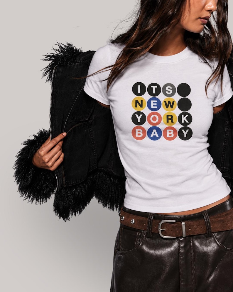 Puede incluir: Una camiseta blanca de manga corta con un estampado gr&aacute;fico de cuatro filas de c&iacute;rculos. Los c&iacute;rculos contienen letras que forman la frase ITS NEW YORK BABY en colores negro, amarillo, rojo y azul, imitando la est&eacute;tica de un letrero de metro.