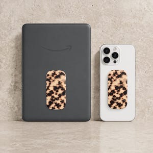 Może przedstawiać: Ciemnoszary czytnik e-booków i biały smartfon, każdy z dołączonym uchwytem do telefonu z wzorem skorupy żółwia. Czytnik e-booków ma logo Amazon. Uchwyty są prostokątne z powtarzającym się wzorem brązu i kremu.