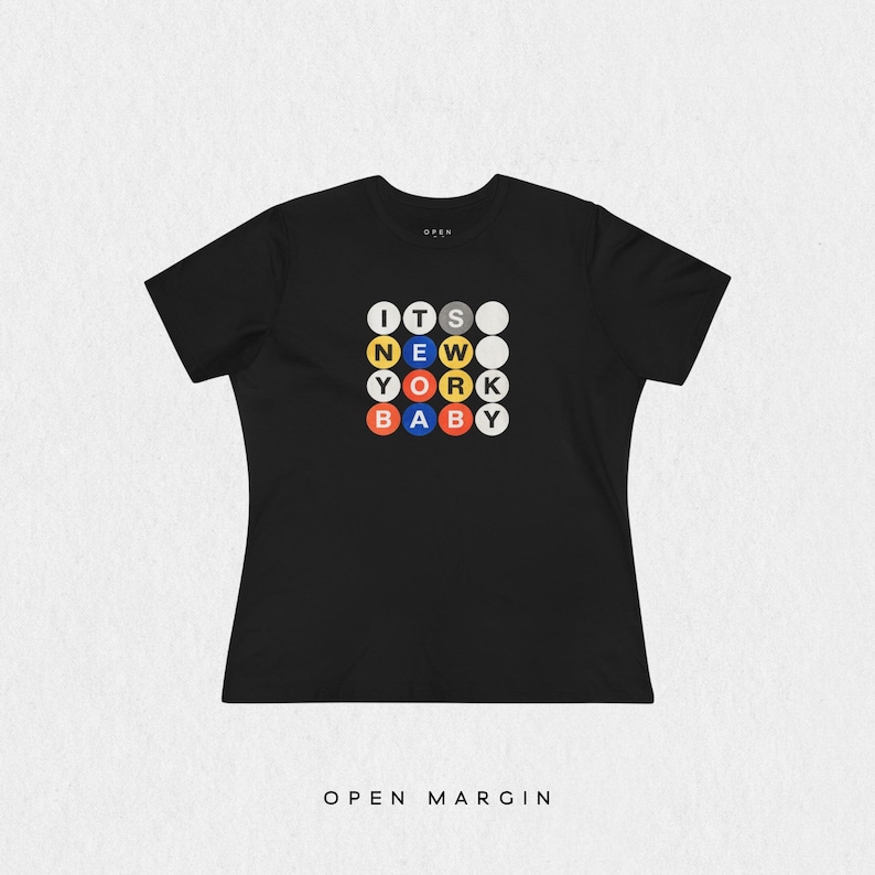 Puede incluir: Una camiseta negra de manga corta con un dise&ntilde;o gr&aacute;fico de c&iacute;rculos de colores dispuestos en una cuadr&iacute;cula. Los c&iacute;rculos contienen letras que forman la frase ITS NEW YORK BABY en una fuente multicolor. La camiseta tiene un cuello redondo simple.
