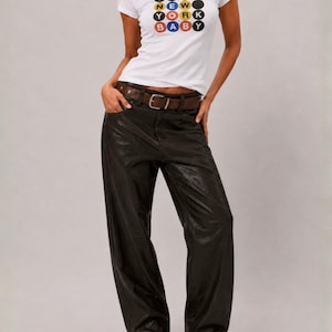 Puede incluir: Una camiseta blanca de manga corta con un gr&aacute;fico de c&iacute;rculos coloridos que contienen el texto ITS NEW YORK BABY, combinada con pantalones negros anchos, un cintur&oacute;n de cuero marr&oacute;n con hebilla de metal plateado y zapatos negros puntiagudos.