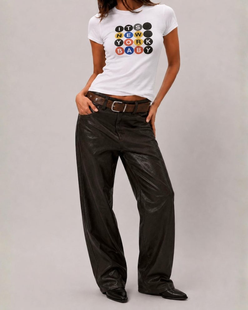 Puede incluir: Una camiseta blanca de manga corta con un gr&aacute;fico de c&iacute;rculos coloridos que contienen el texto ITS NEW YORK BABY, combinada con pantalones negros anchos, un cintur&oacute;n de cuero marr&oacute;n con hebilla de metal plateado y zapatos negros puntiagudos.