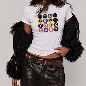 Puede incluir: Una camiseta blanca de manga corta con una cuadr&iacute;cula de c&iacute;rculos de colores que contienen el texto "ITS NEW YORK BABY" en negro, amarillo, rojo y azul. La camiseta se combina con pantalones negros de efecto piel y un cintur&oacute;n de ante marr&oacute;n con tachuelas plateadas.