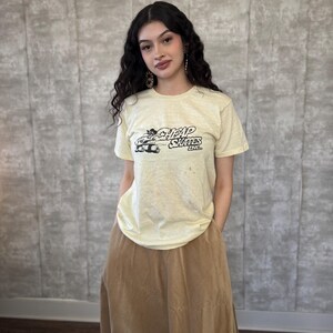 May include: A woman wearing a pale yellow t-shirt with a black and white graphic of a cartoon character and the text "Cheap Skates Inc." The t-shirt is paired with a brown skirt.
