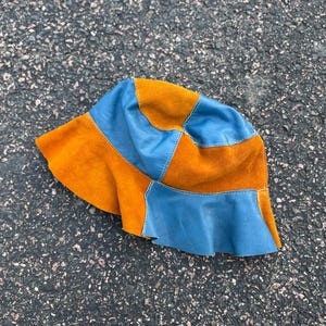 May include: A patchwork bucket hat in shades of orange and blue. The hat features a combination of suede and leather panels, with a slightly ruffled brim. The hat is designed with a classic bucket hat silhouette.