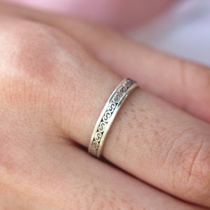 May include: A silver ring with a delicate, intricate design. The ring is worn on a person's finger.