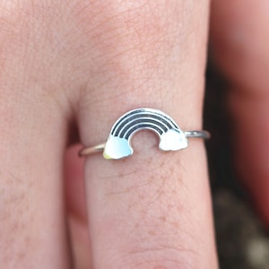 May include: A silver ring featuring a rainbow design. The rainbow has black stripes and white clouds at each end. The ring is worn on a finger, showcasing the jewelry's delicate details and craftsmanship.