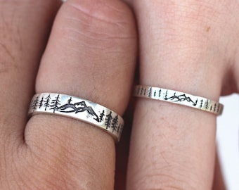 May include: Two silver rings with a nature-inspired design. One ring features a mountain range and trees, while the other ring features a row of trees.