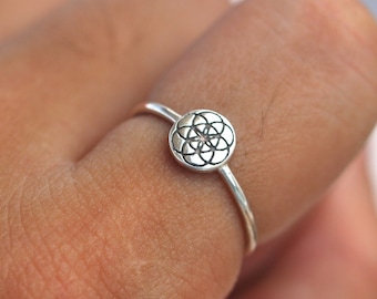Op de afbeelding: Een zilveren ring met een kleine, ronde, zilveren bedel met een geometrisch ontwerp. De ring zit aan een vinger.