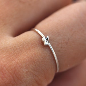 May include: A delicate silver ring featuring a small, double heart design. The ring is worn on a finger.