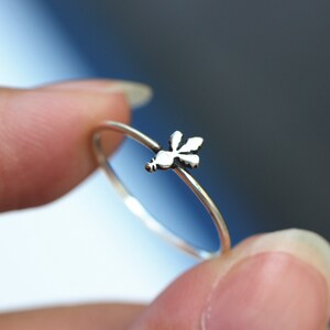 May include: A delicate silver ring featuring a small dragonfly design. The ring's band is thin and simple, with the dragonfly charm positioned at the top. The ring is held between two fingers against a blurred background.