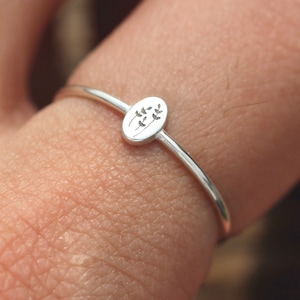 May include: A delicate silver ring with a small oval-shaped design featuring a floral motif. The ring is worn on a finger.
