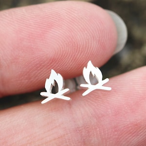 Puede incluir: Un par de pendientes de plata con un diseño de fogata. Los pendientes son pequeños y delicados, y serían un gran regalo para cualquier persona que ame acampar o el aire libre.