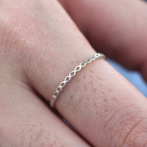 May include: A delicate silver ring with a repeating infinity symbol pattern. The ring is thin and sits on a finger, highlighting its intricate design. The ring's design is simple yet elegant, suitable for everyday wear.