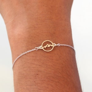 May include: A silver chain bracelet with a gold circle charm featuring a mountain silhouette.
