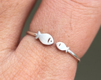 May include: A silver ring with two fish facing each other. The ring is on a person's finger.