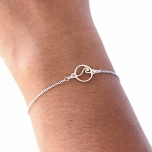 Pode incluir: Um bracelete de corrente de prata com um pequeno charme circular com um design de onda.