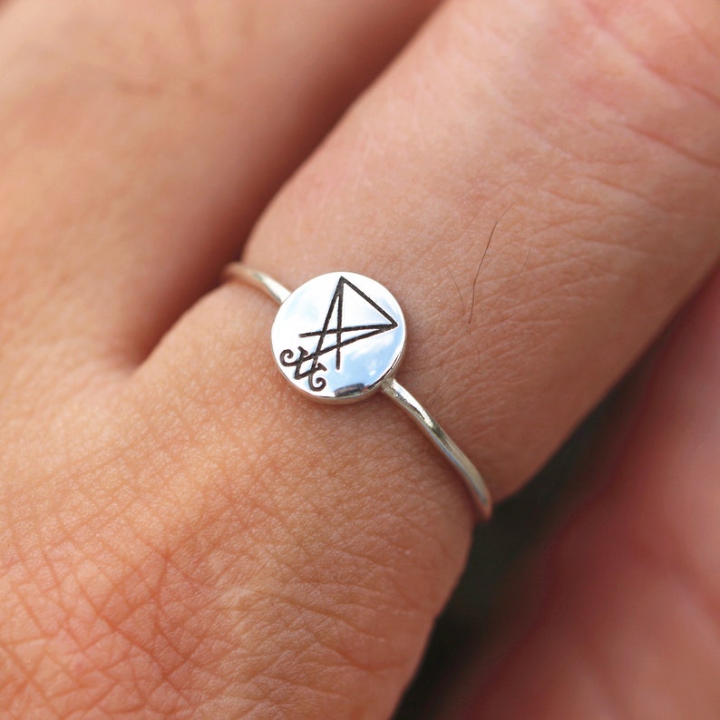 May include: A silver ring with a circular design featuring a triangle and a symbol inside. The ring is worn on a finger.